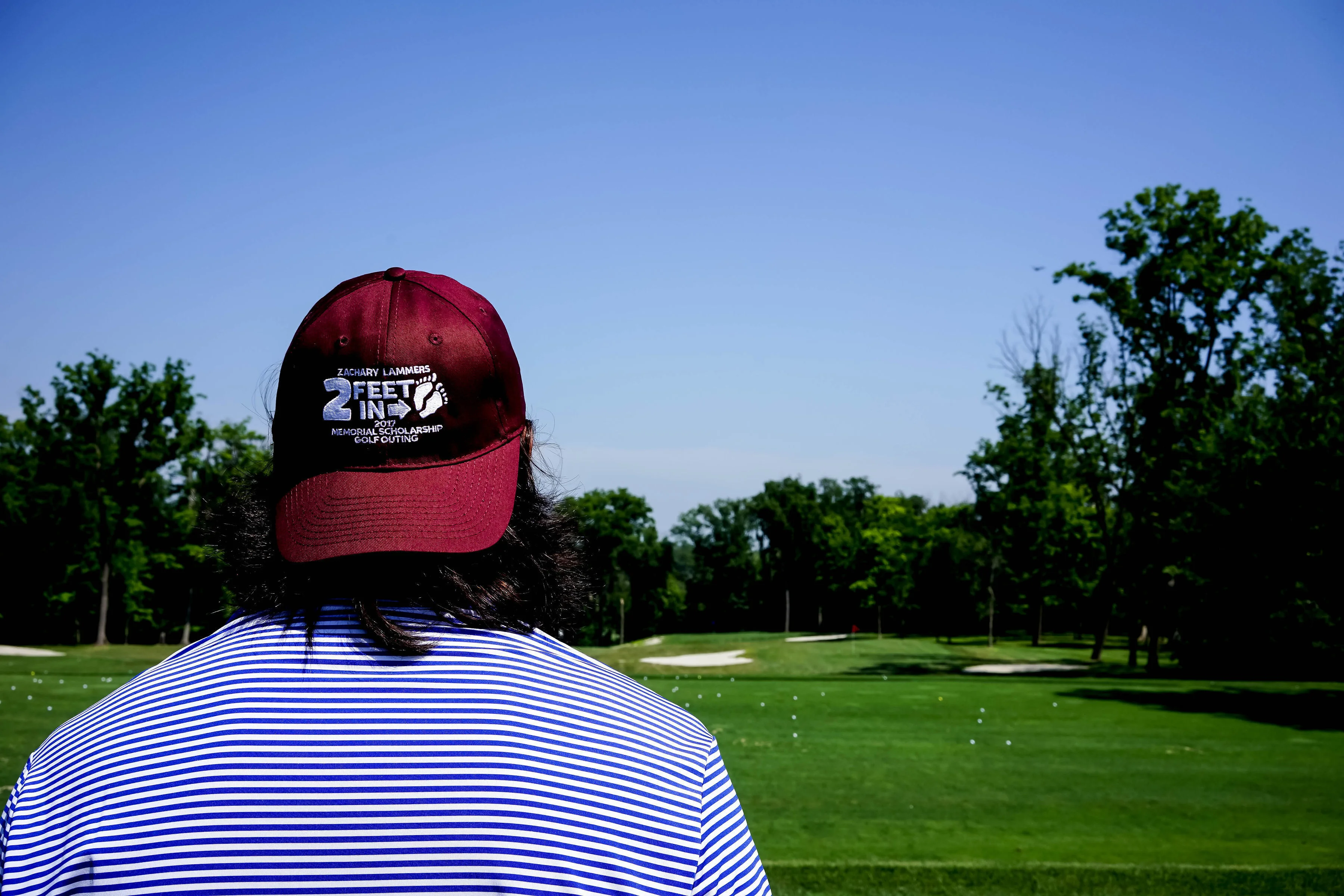 Person wearing a maroon Zachary Lammers Memorial Scholarship Golf Outing cap, viewed from behind, looking out over a sunlit golf course.