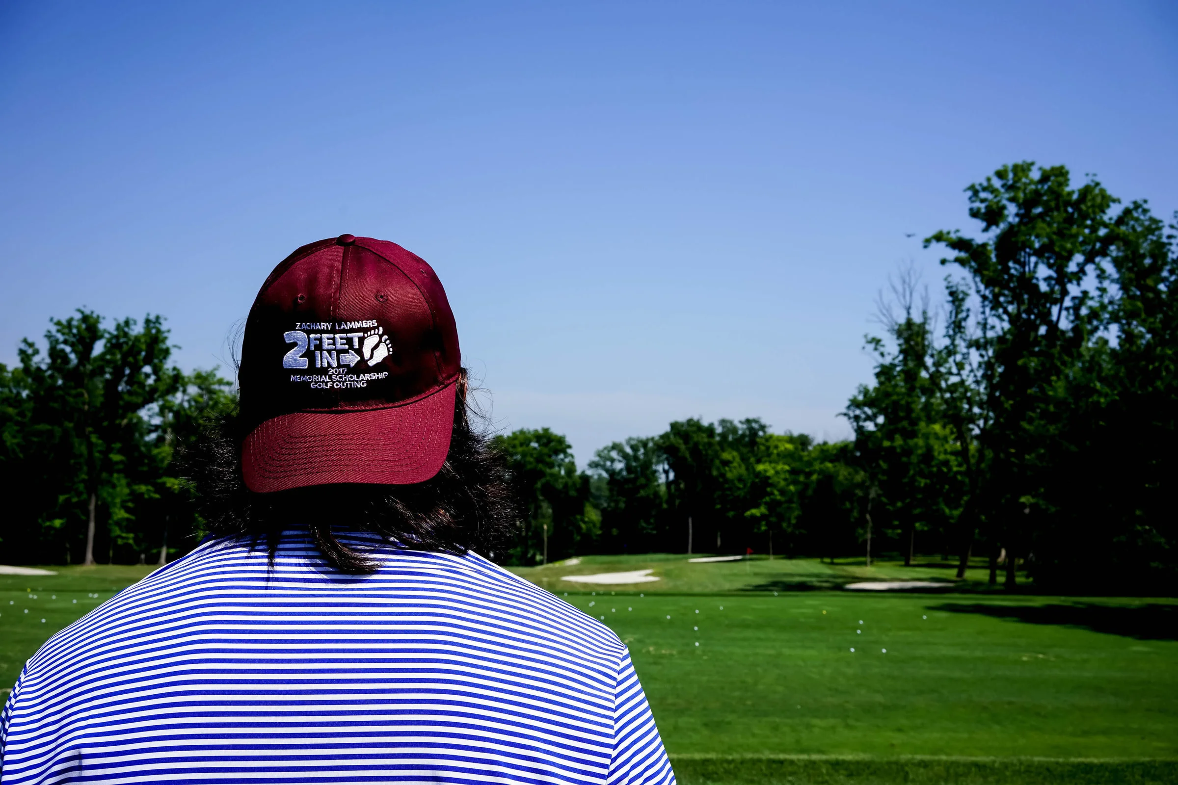 Person wearing a maroon Zachary Lammers Memorial Scholarship Golf Outing cap, viewed from behind, looking out over a sunlit golf course.