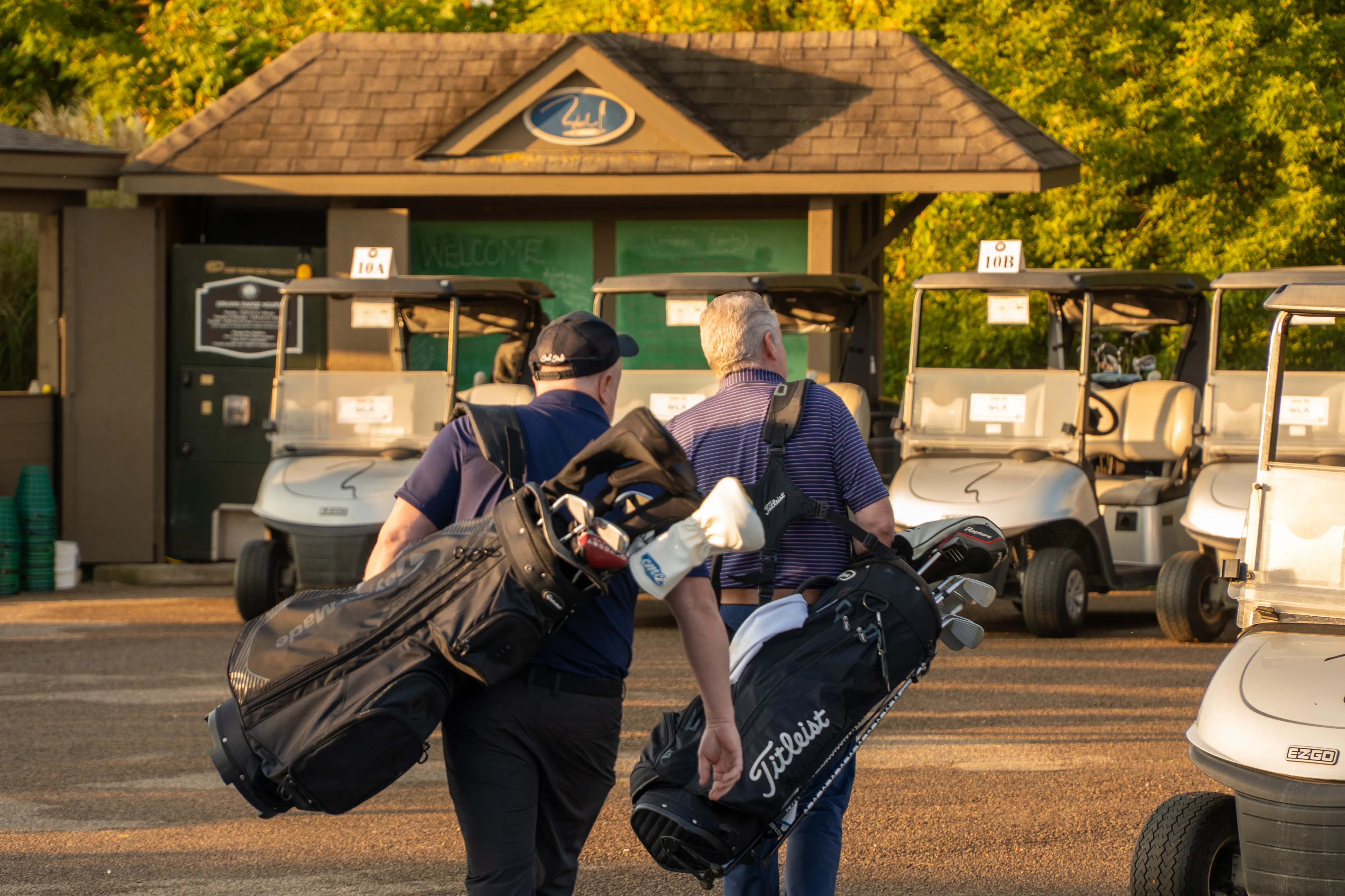 Two golfers carrying their bags toward a row of carts at a tournament staging area.