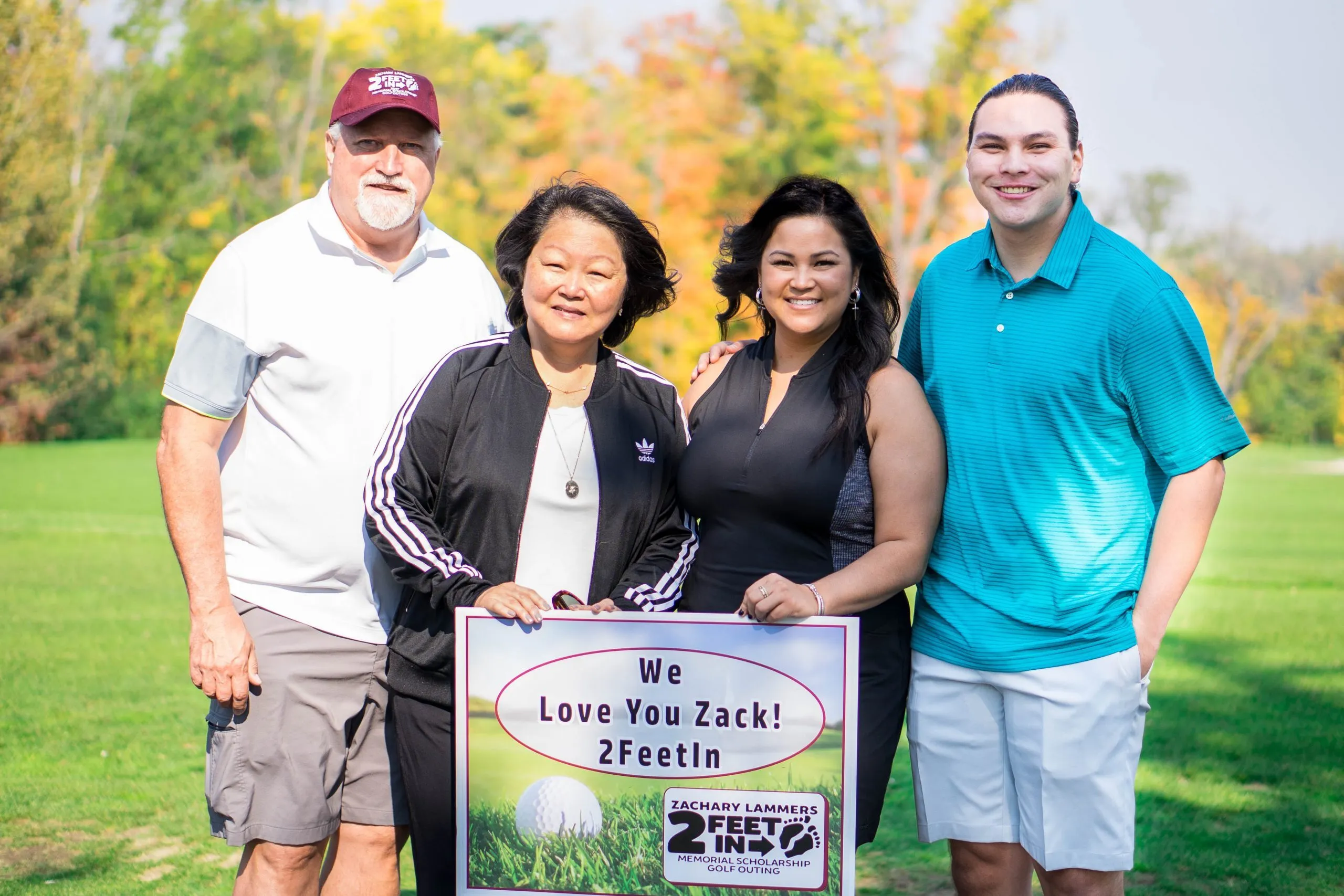 Tom and Gabe Lammers with supporters on a golf course holding a memorial sign reading We Love You Zack! 2FeetIn for the Zachary Lammers Memorial Scholarship Golf Outing.