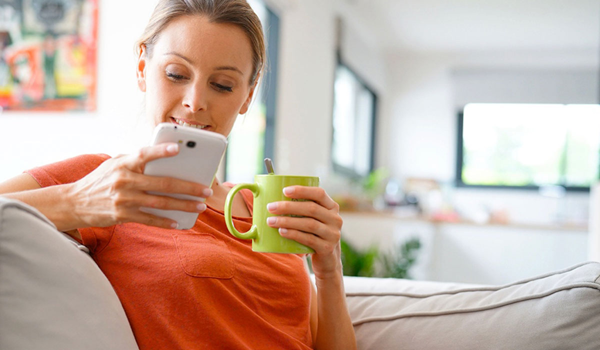 Woman wearing orange shirt sitting on couch holding phone and green coffee mug