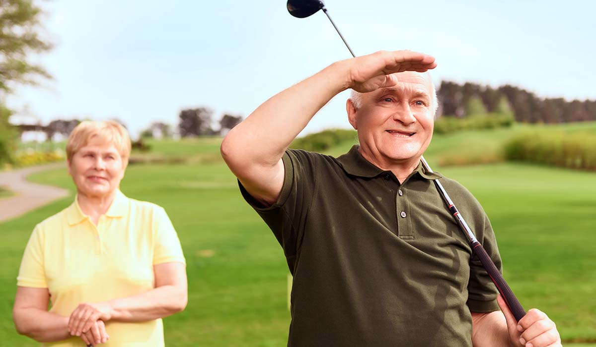 Elderly male golfer watching ball in golf course with woman wearing yellow in the background